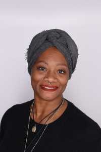 Picture of Natalie Thomas, Program Coordinator of  Black Programs. The image is a portrait of a person against a plain, light-colored background. The person is smiling and looking directly at the camera. They are wearing a dark gray headwrap, which is intricately tied at the front. Their skin has a warm tone, and they have neatly groomed eyebrows and full, red lips. They have a small nose piercing on the right nostril and a septum ring. They are dressed in a black top and are wearing two necklaces: a thin silver chain and a black cord, both of which have circular pendants.